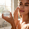 Woman applying facial cream with a jar of the product in a bathroom setting
