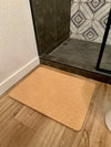 Beige cork bathroom mat on a wooden floor in front of a glass shower door.