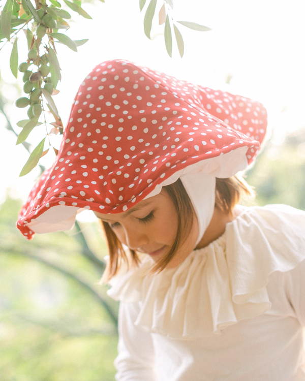Person wearing a red polka dot bonnet and white blouse outdoors with greenery in the background