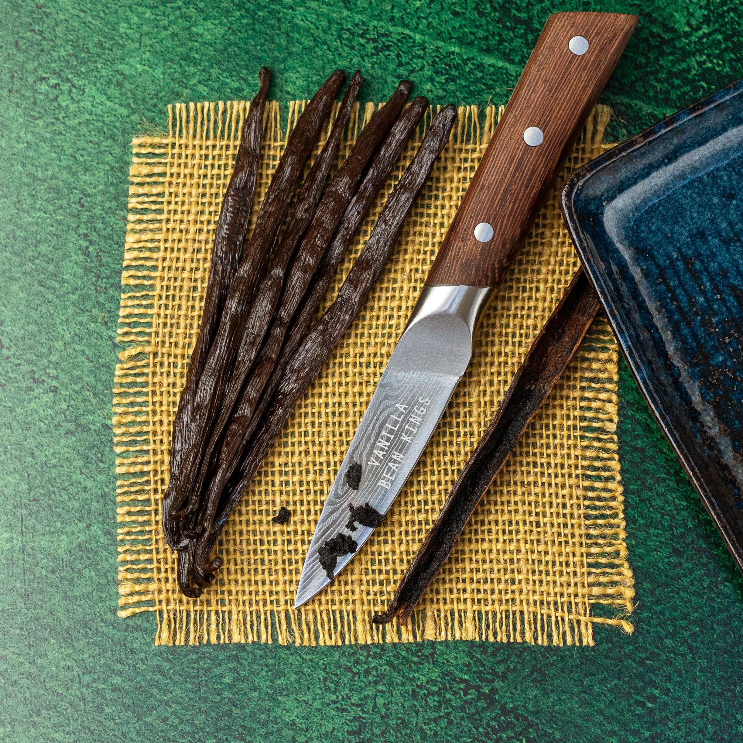 Vanilla beans with a knife on a yellow cloth against a green background