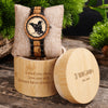 Wooden watch with engraved design on a decorative stand, surrounded by greenery.