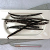 Vanilla beans on a white plate with a metal tool on a gray surface