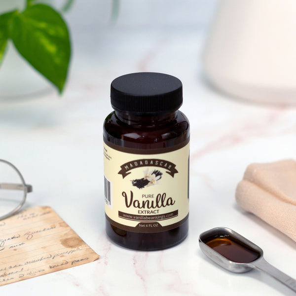 Bottle of vanilla extract on a surface with a spoonful of extract and a plant in the background.