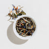 White bowl filled with loose leaf White Coconut Crème tea showing coconut pieces, safflower, and cornflower petals on a light gray background
