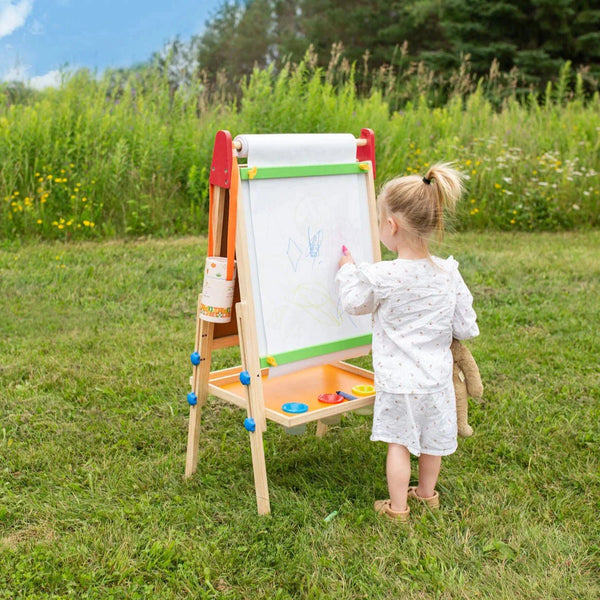 Child drawing on an outdoor easel in a grassy area with trees in the background