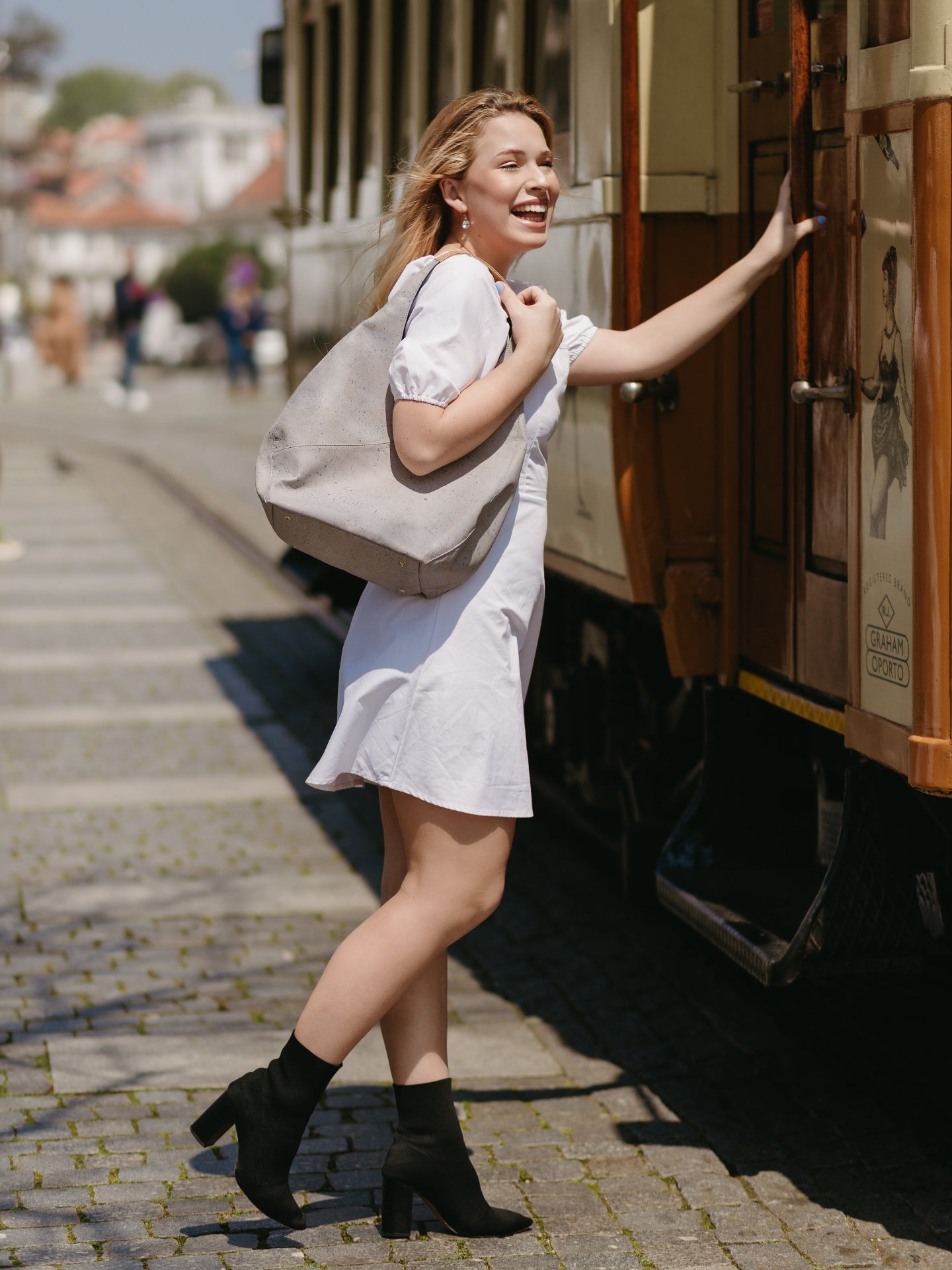Woman in a white dress with a beige bag standing next to a vintage tram on a city street.