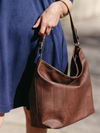 Brown leather handbag held by a person wearing a blue dress.