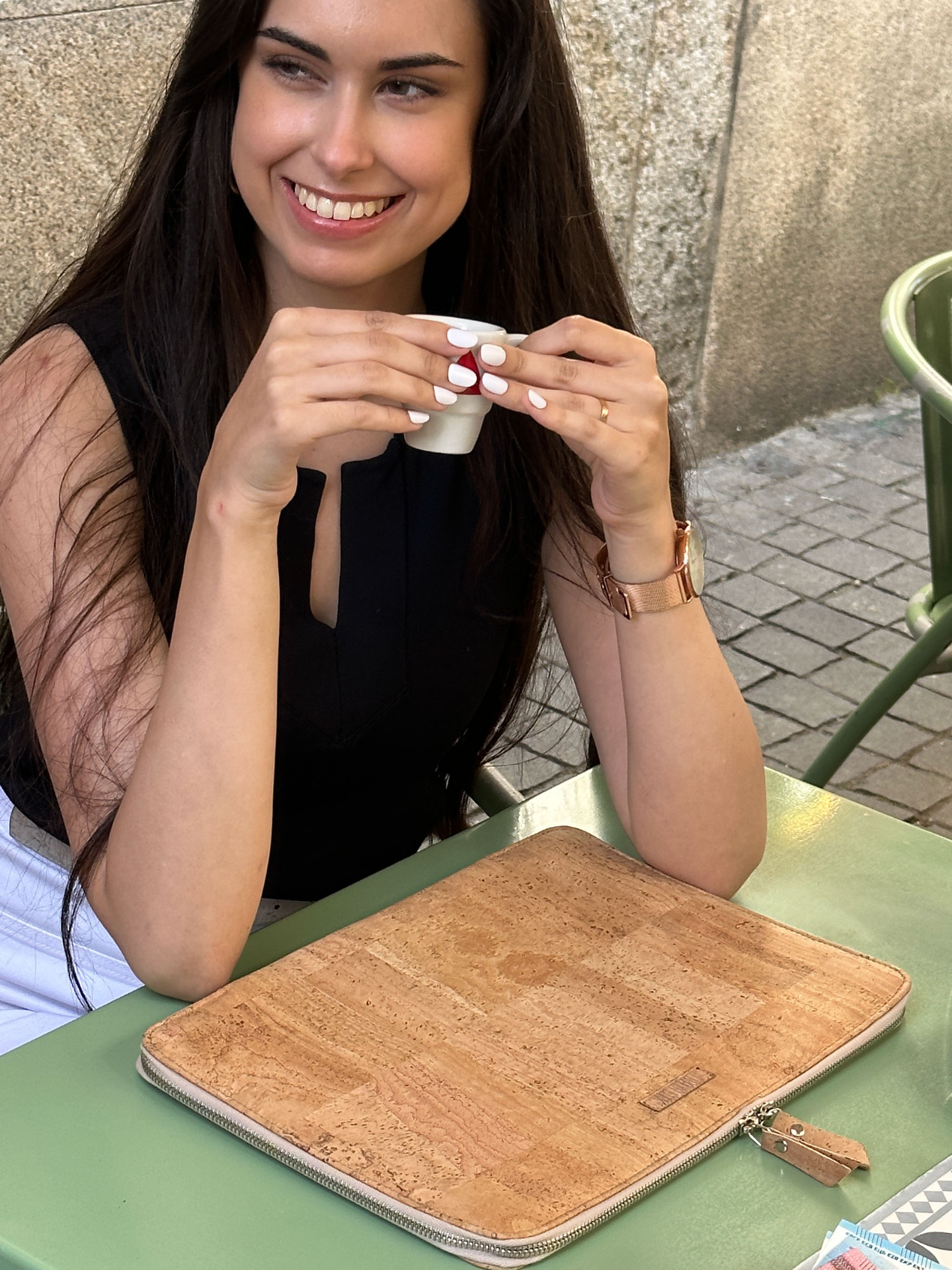Woman holding a cup outdoors with a cork laptop sleeve on a table in front of her.