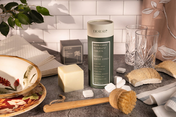 Eco-friendly bamboo dish scrubbing tool alongside sustainable kitchen cleaning products including solid dish soap block and plant cellulose sponge
