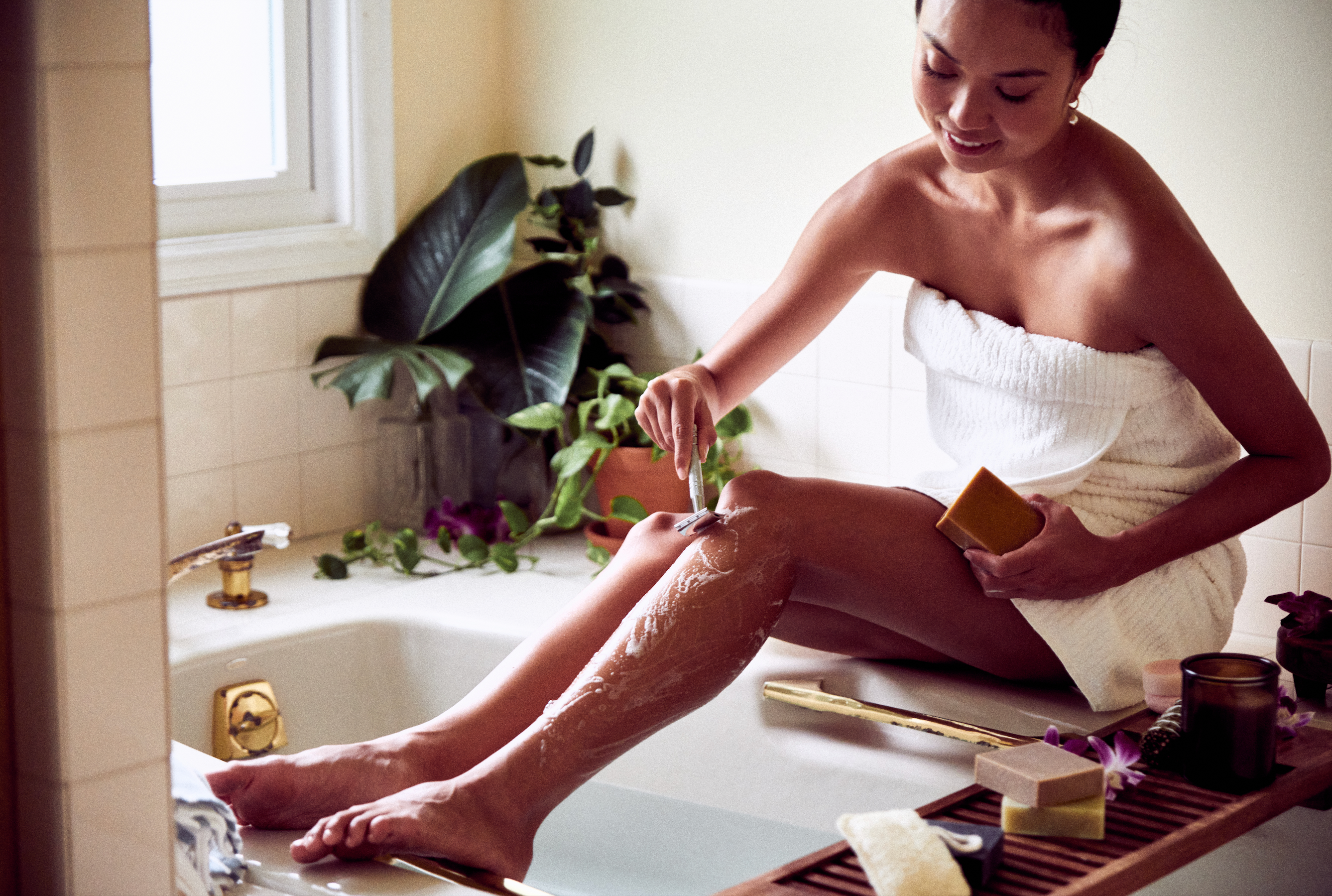 Woman using Desesh metal safety razor and solid shave soap beside a bathtub as part of a sustainable self-care routine