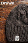 Brown knit beanie with a brand logo on a wooden surface, labeled 'Brown'.