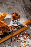 Jar of Fine & Raw chocolate hazelnut butter on a wooden board with bread and nuts.