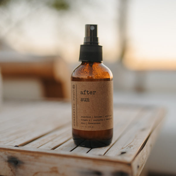 A bottle of 'After Sun' spray with a spray nozzle, placed on a surface alongside a pair of sunglasses and a cloth, under natural lighting.
