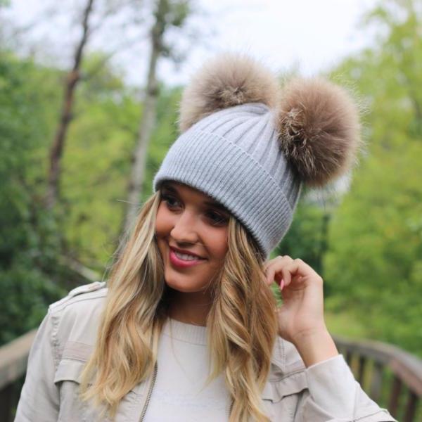 Woman wearing a light gray knit beanie with two large pom-poms outdoors.