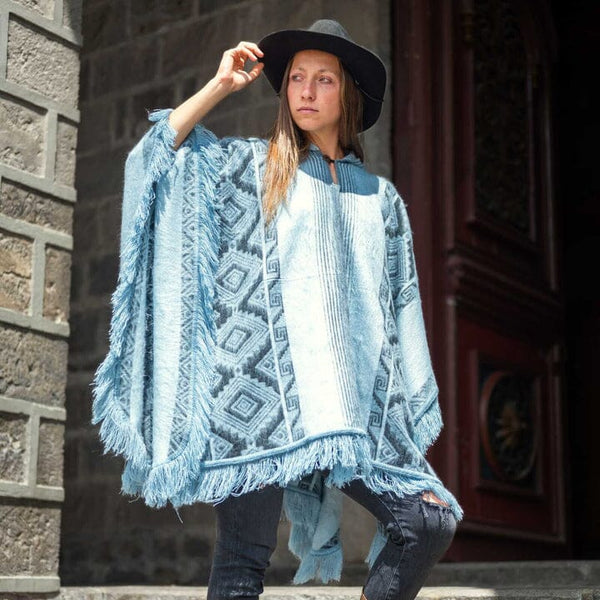 Woman wearing a blue patterned poncho and black hat against a stone wall.