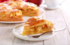 Slice of apple pie on a plate with a fork, surrounded by more pies and apples on a wooden table.