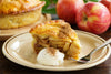 Slice of apple pie on a plate with whipped cream and a fork, surrounded by apples.