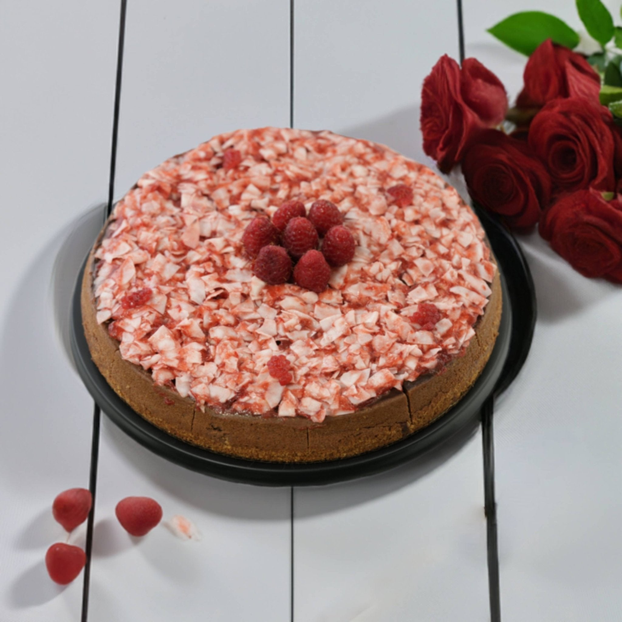 Round cake with pink crumb topping and raspberries on a white wooden surface with red roses.
