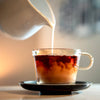 Glass cup being filled with a creamy liquid from a white pitcher on a neutral background