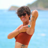 Woman applying sunscreen on a beach with a clear blue sky and ocean in the background. NANAH Rosemary natural deodorant — aluminum-free, 24-hour protection for excessive sweating. Magnesium, zinc ricinoleate & rosemary oil. Plastic-free. Shop now.