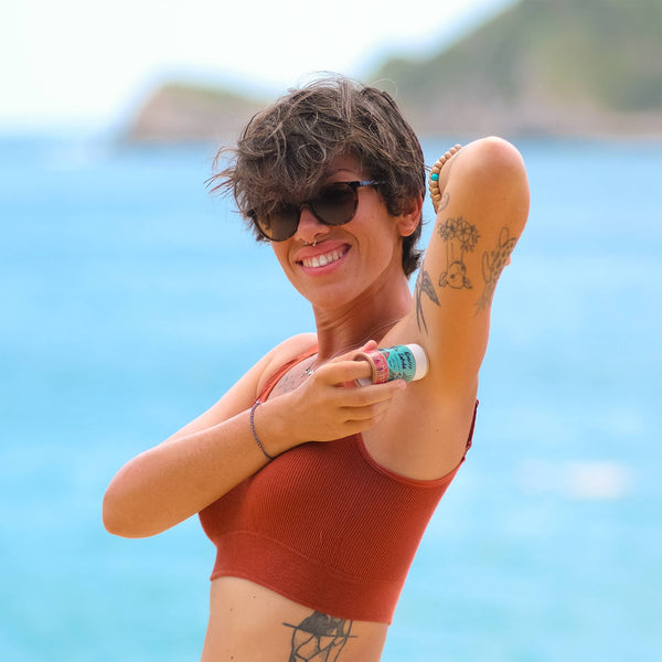 Woman applying sunscreen on a beach with a clear blue sky and ocean in the background. NANAH Rosemary natural deodorant — aluminum-free, 24-hour protection for excessive sweating. Magnesium, zinc ricinoleate & rosemary oil. Plastic-free. Shop now.