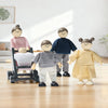 Four wooden toy children figures with fabric clothing and black feet, including a stroller figure, arranged on a light wood floor against a softly blurred indoor background.