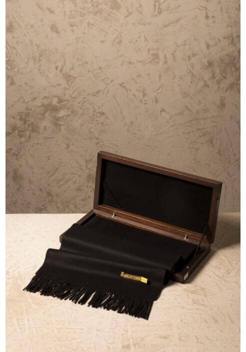 Black scarf with fringes draped over a wooden box against a textured beige wall.