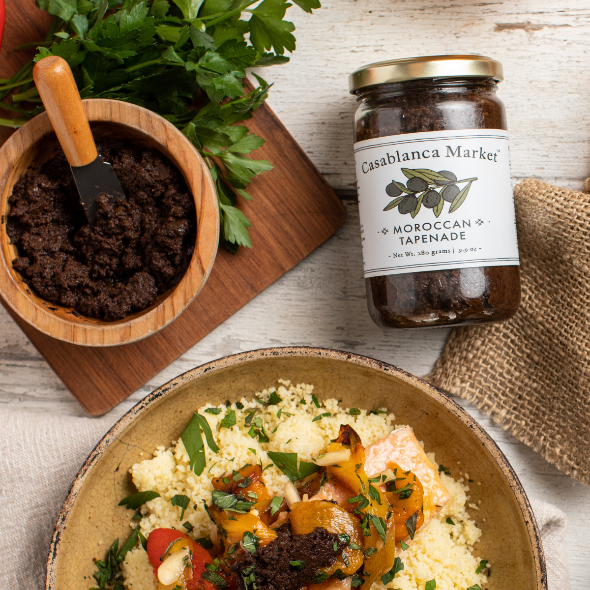 Jar of Casablanca Market Moroccan Tapenade next to a bowl of couscous with vegetables on a wooden surface.