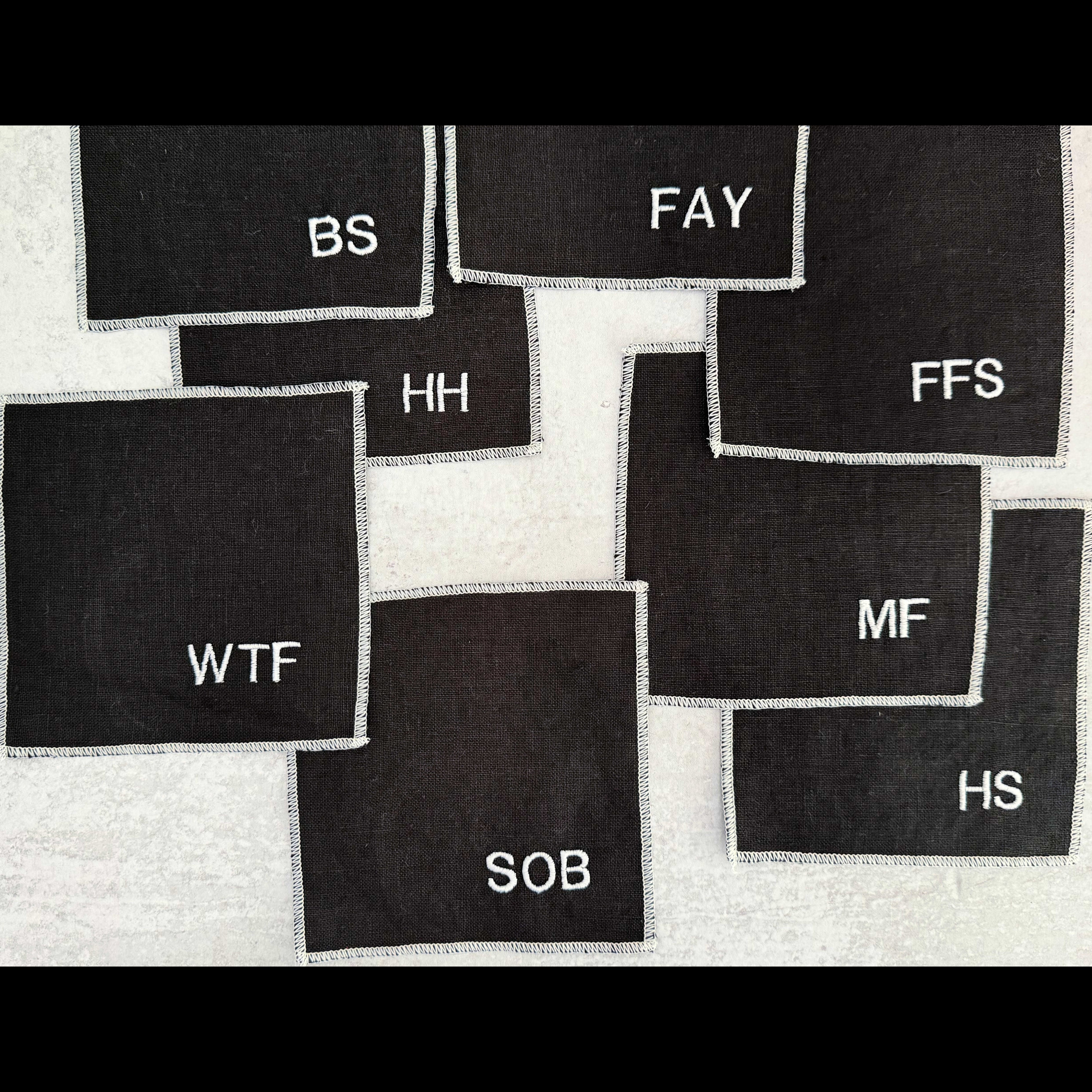 Set of eight black linen sweary cocktail coasters with white letter monogram design, laid flat on a surface