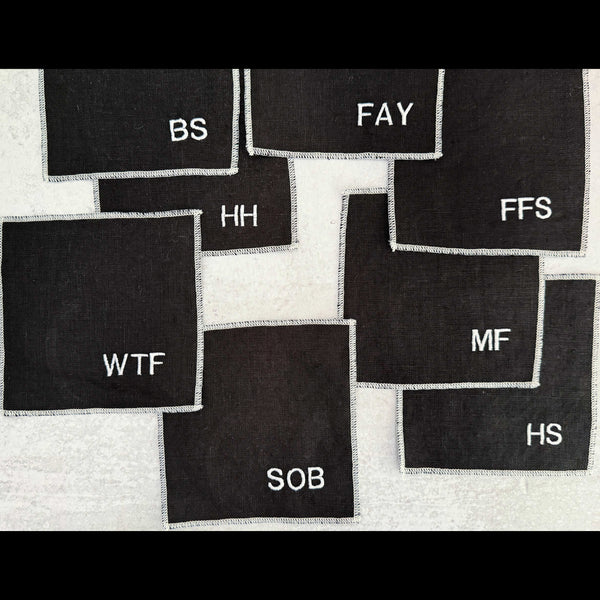 Set of eight black linen sweary cocktail coasters with white letter monogram design, laid flat on a surface