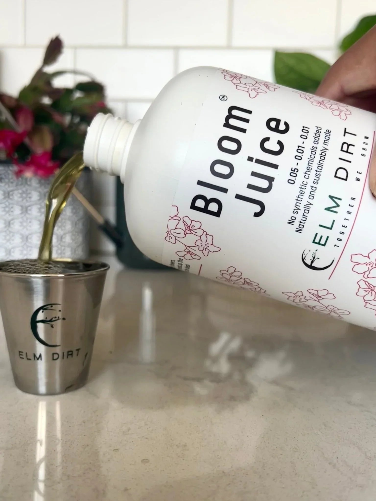 Bottle of 'Bloom Juice' being poured into a metal cup on a kitchen counter. Bloom Power Bundle — fast-acting Bloom Juice with 150+ microbes & slow-release Bloomin' Soil for explosive flowering. More blooms, bigger color. Perfect garden gift.