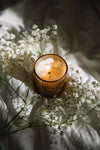Candle in a textured holder surrounded by delicate white flowers on a soft surface