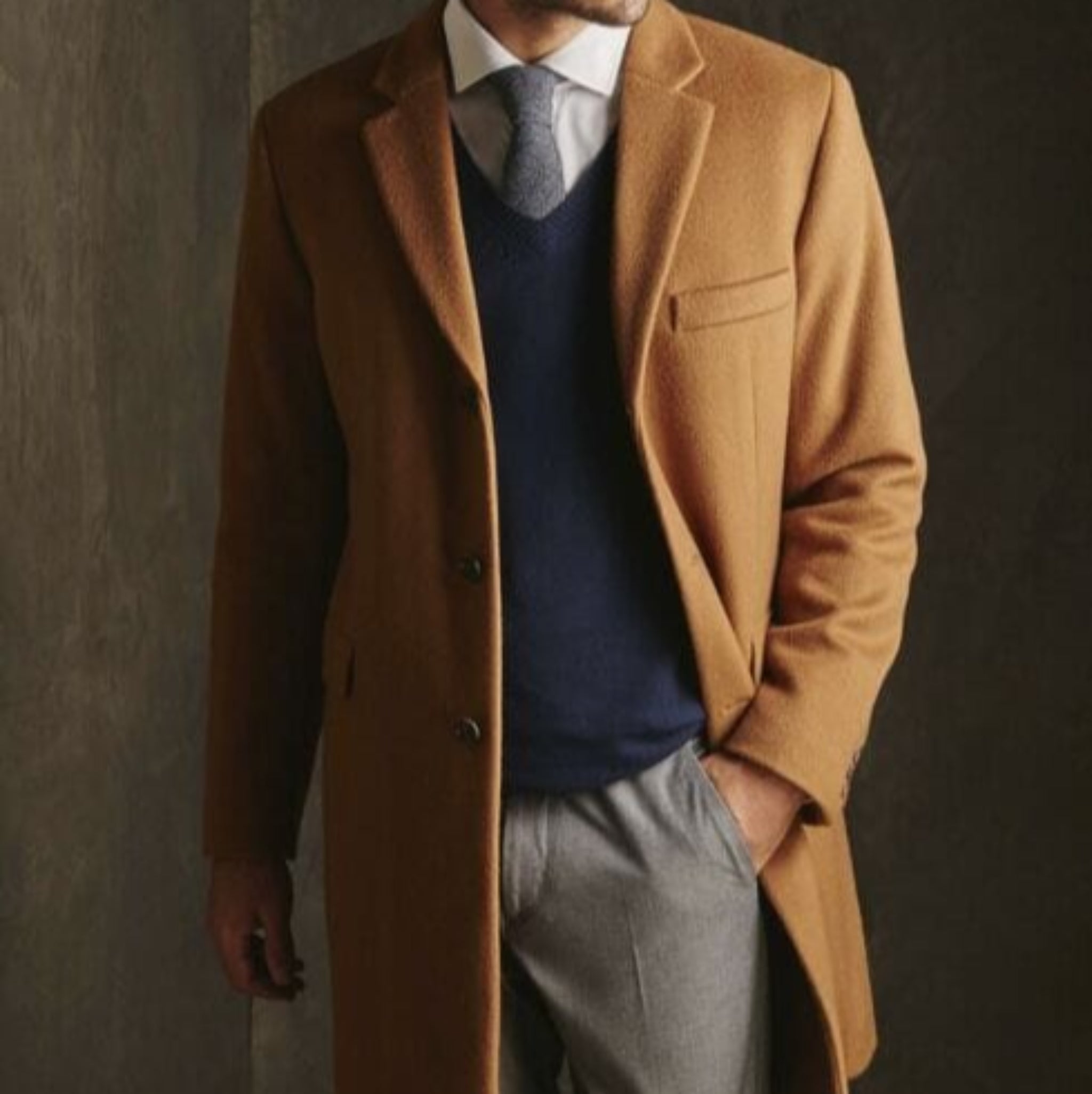 Man wearing a brown coat over a blue sweater and gray pants against a dark wall.