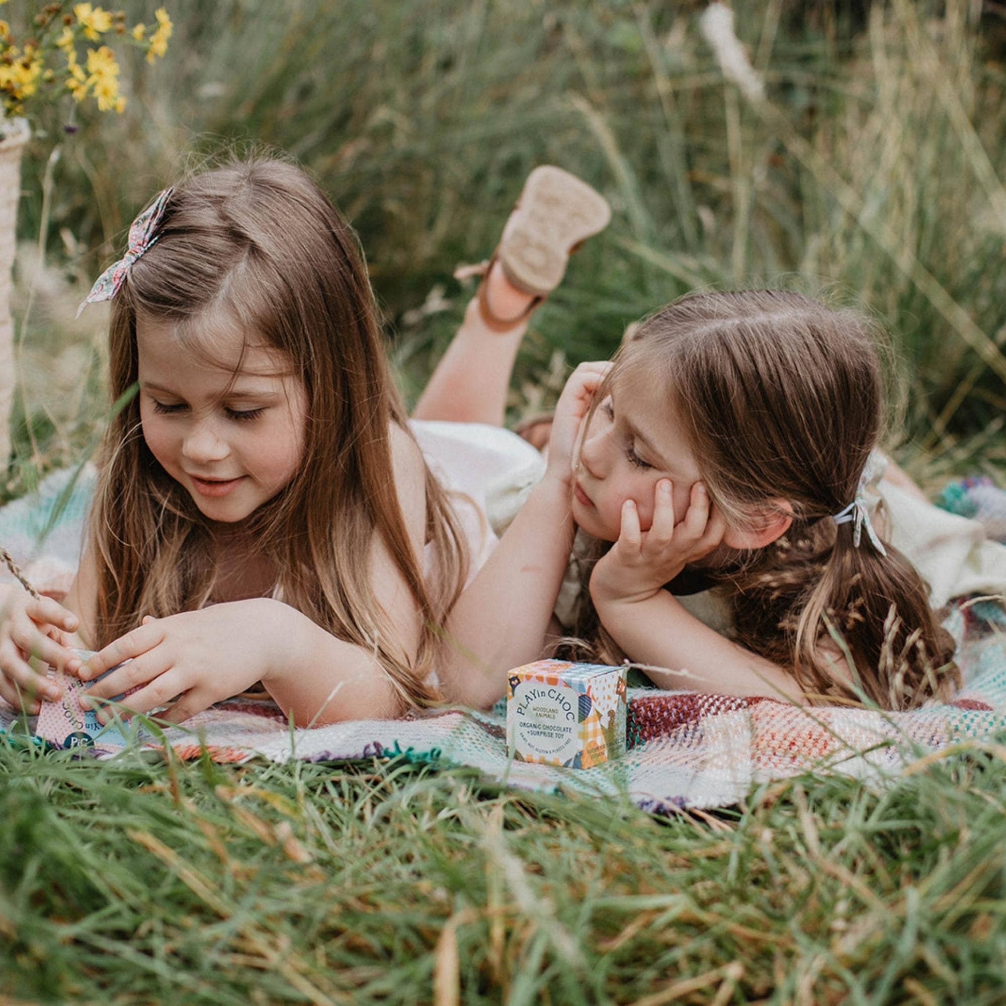 Two young girls lying on a blanket in a grassy field with a small container of cream.