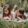 Two young girls lying on a blanket in a grassy field with a small container of cream.