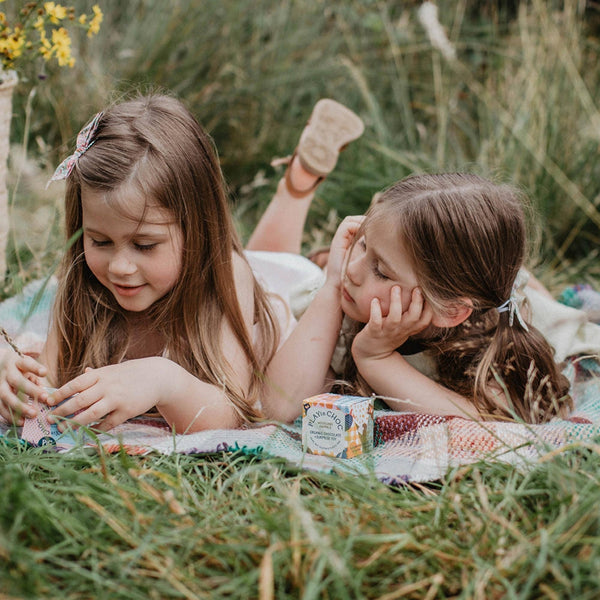 Two young girls lying on a blanket in a grassy field with a small container of cream.