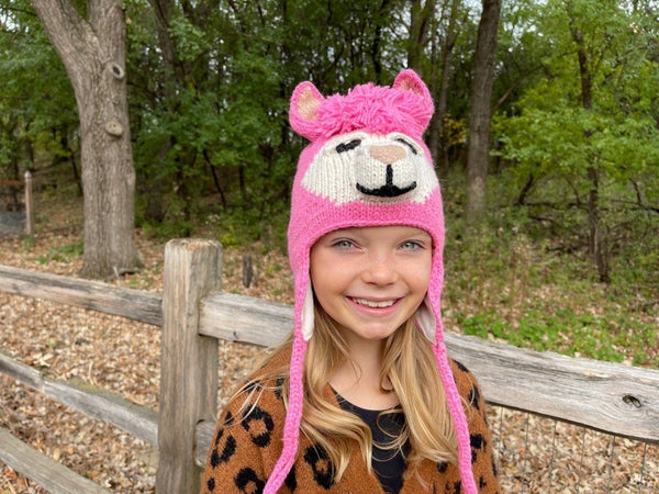 Child wearing a pink animal-themed hat outdoors near a wooden fence.