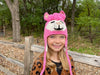 Child wearing a pink animal-themed hat outdoors near a wooden fence.