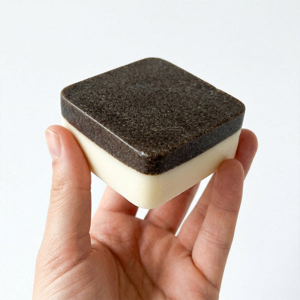 Hand holding a bar of soap with a textured black top and white bottom against a light gray background