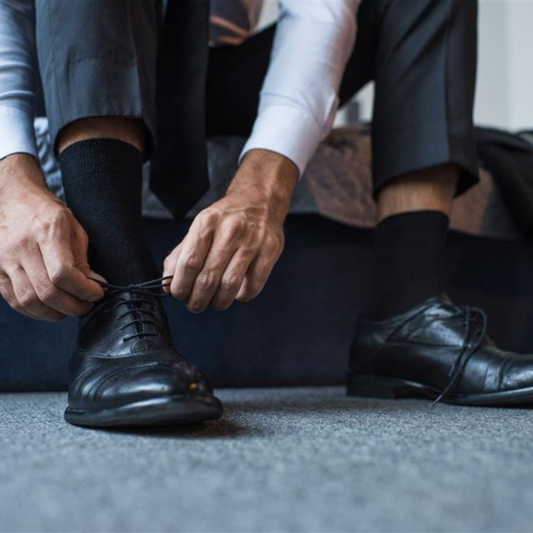 Person tying black shoes on a carpeted floor with a neutral background. Classic Alpaca Socks – soft, breathable, and versatile for year-round wear. Ethically made in Peru. Shop timeless alpaca comfort today.