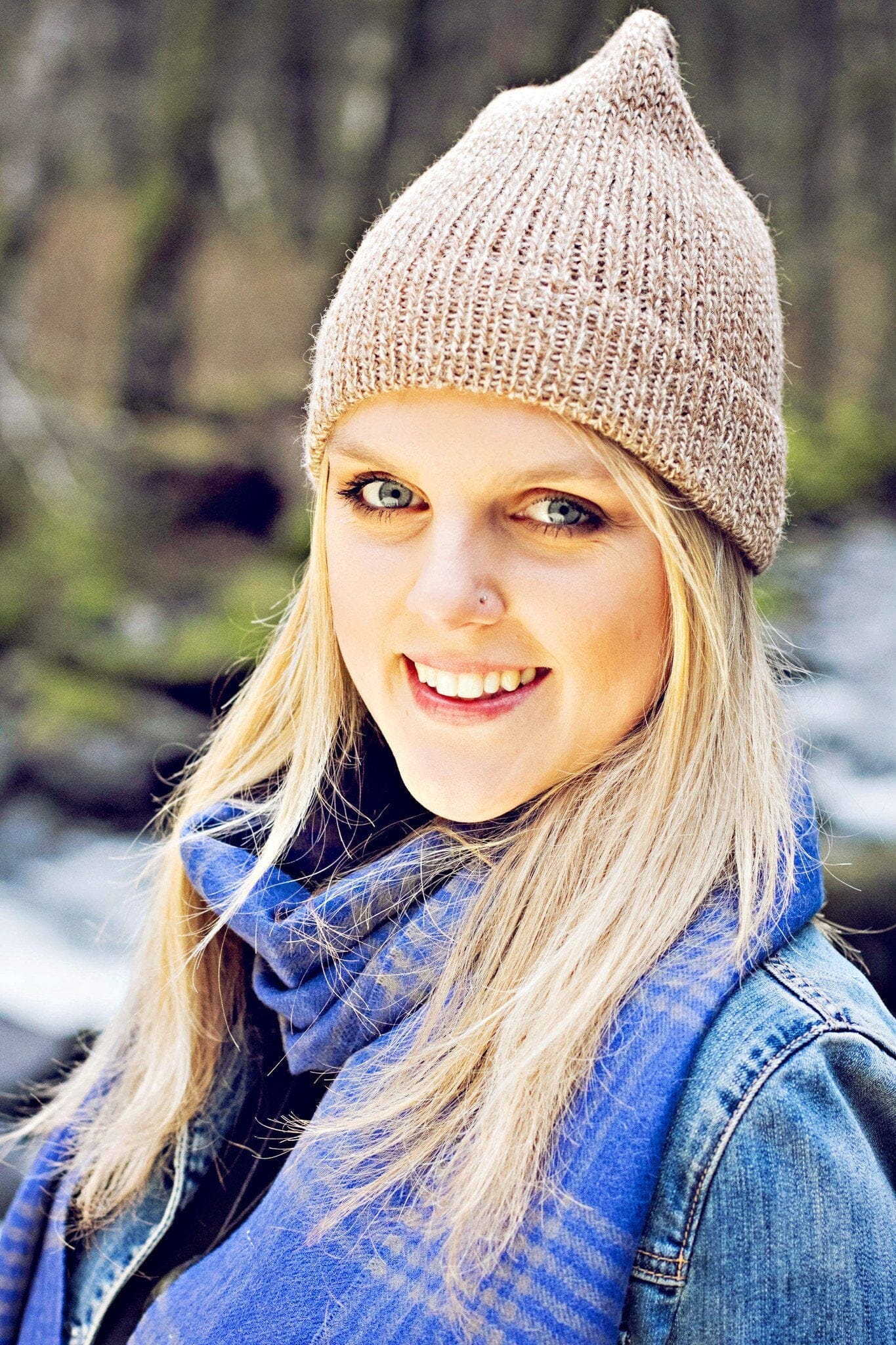 Woman wearing a beige knit hat and blue scarf outdoors with blurred natural background