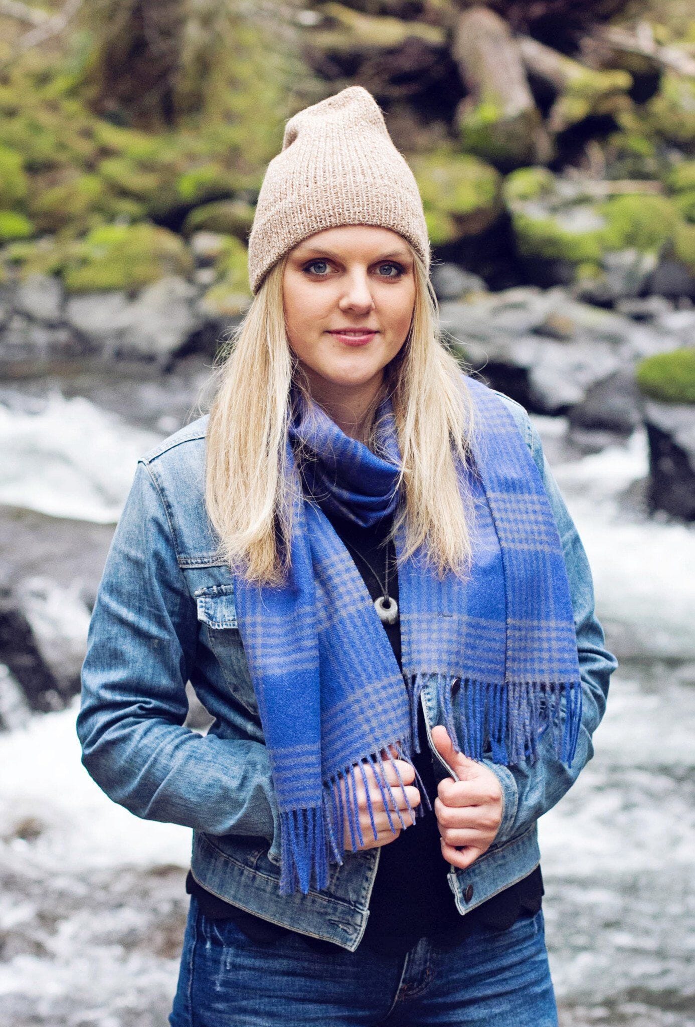 Woman wearing a blue plaid scarf, beige beanie, and denim jacket by a stream.