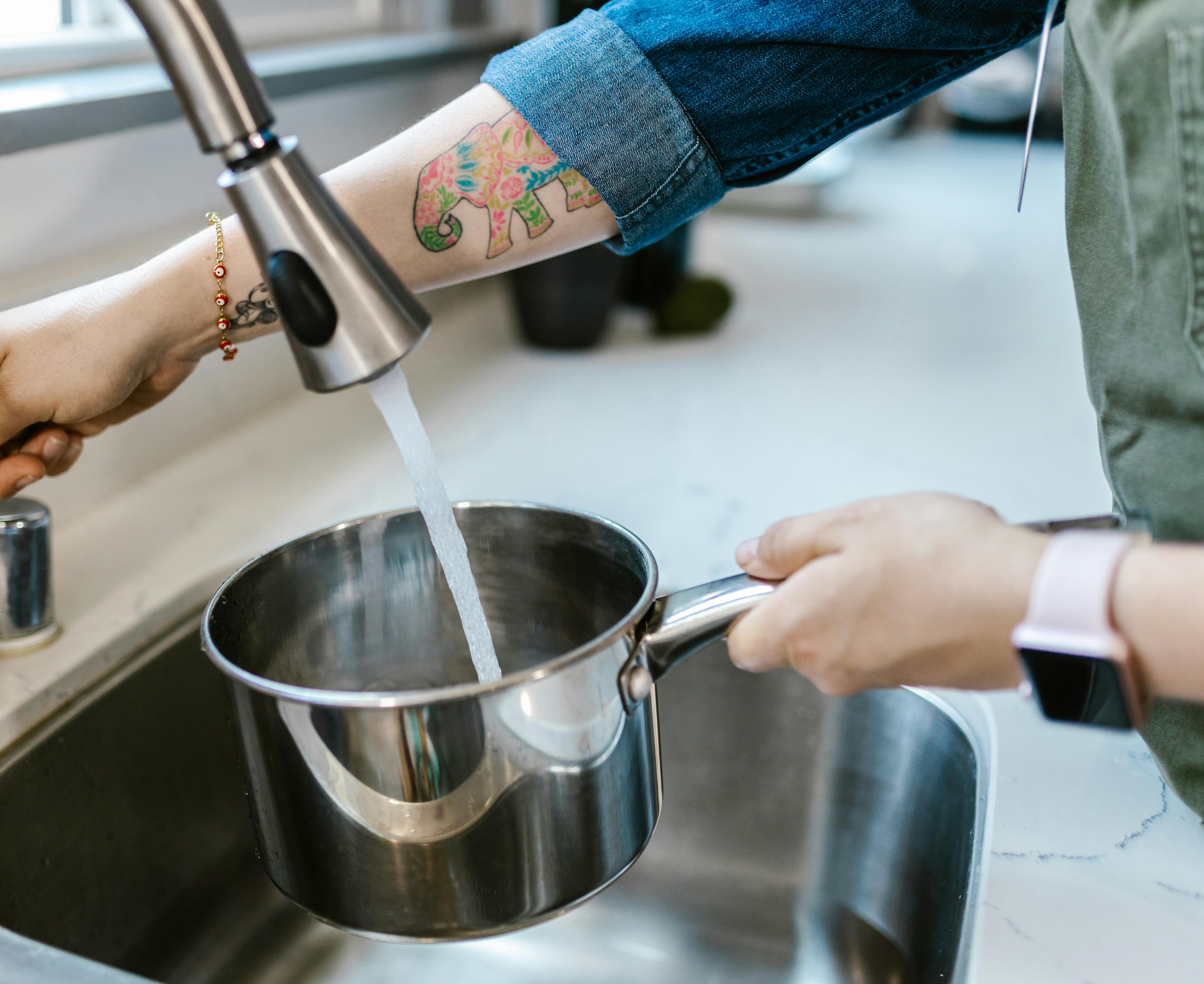 Person filling a metal pot with water from a faucet in a kitchen.