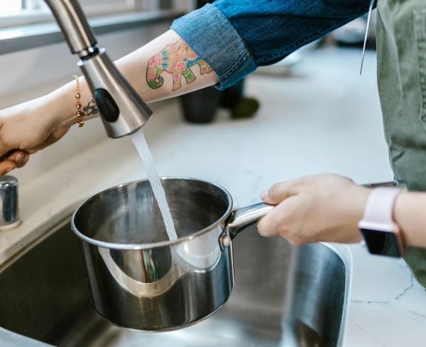 Person filling a metal pot with water from a faucet in a kitchen.