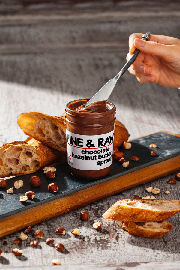 Jar of Fine & Raw chocolate hazelnut butter with a spoon and bread on a wooden board.