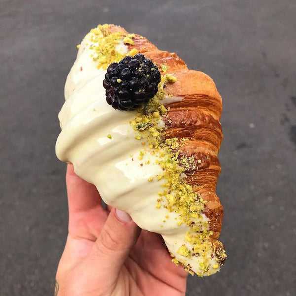 Hand holding a croissant with white cream, blackberry, and pistachio on a dark background