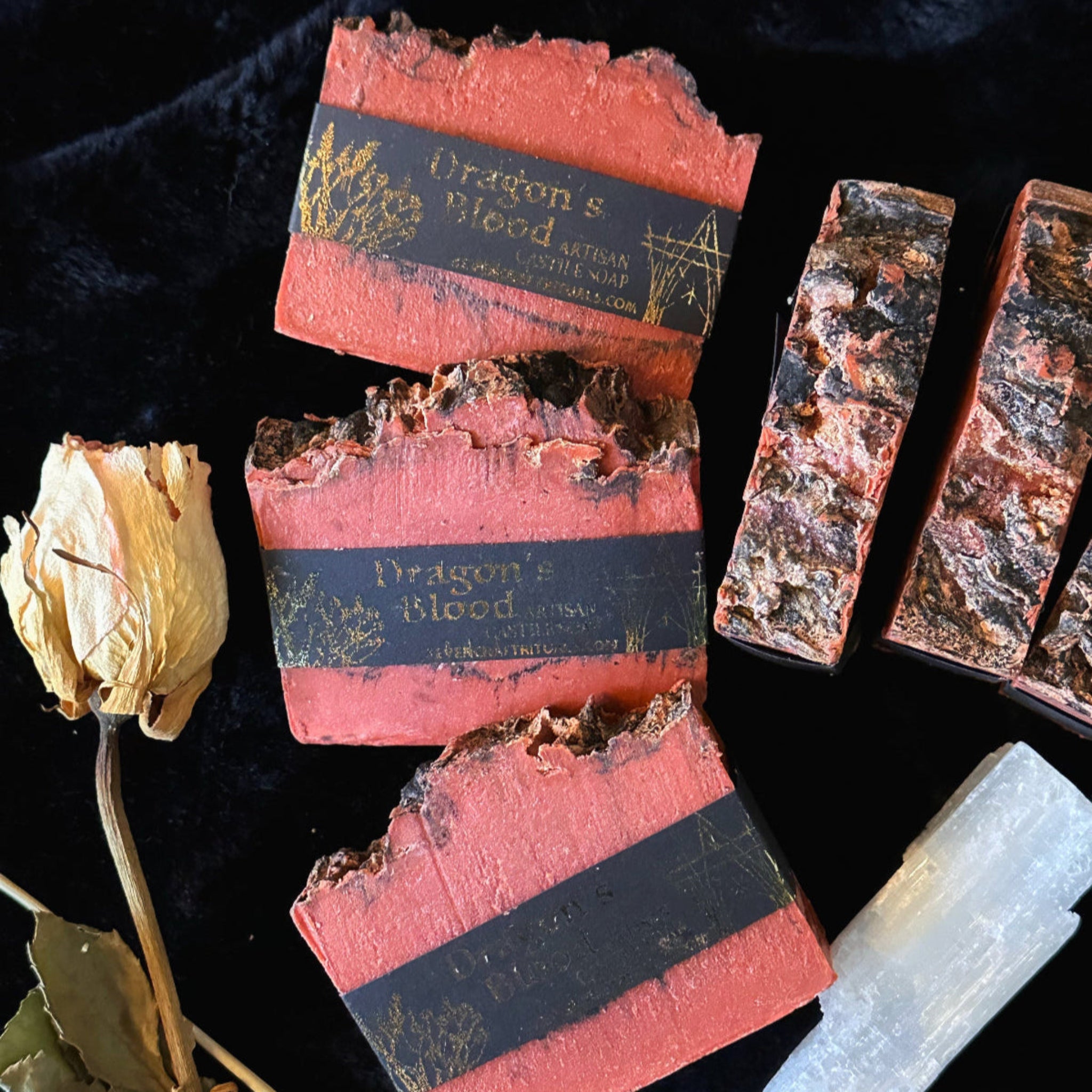 Red soap bars with black labels on a dark background, accompanied by a dried flower and crystal.