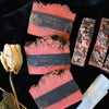 Red soap bars with black labels on a dark background, accompanied by a dried flower and crystal.
