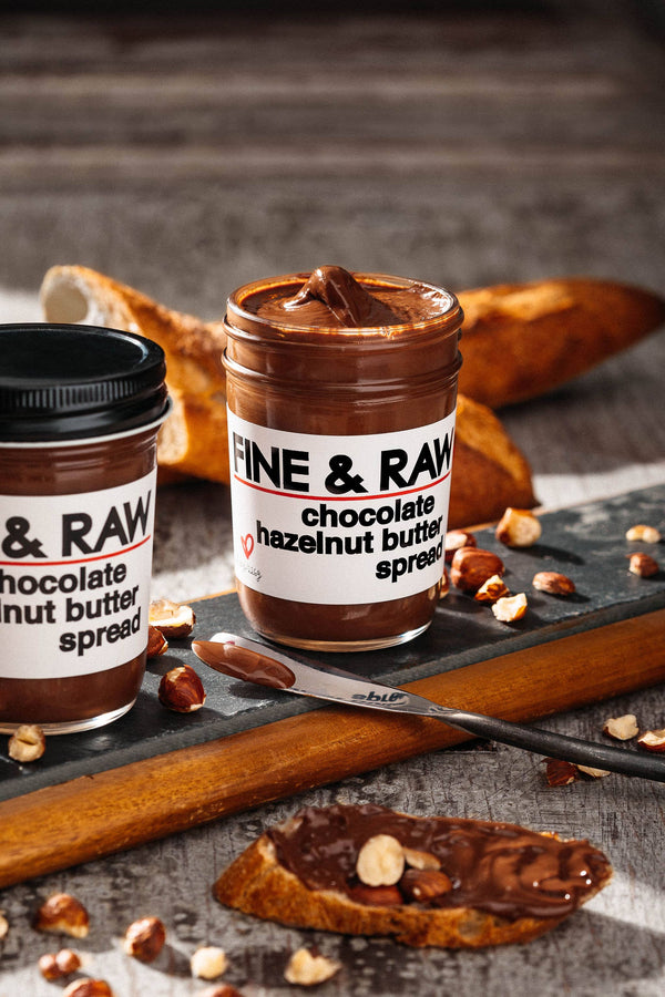 Two jars of Fine & Raw chocolate hazelnut butter spread on a wooden board with a spoon and bread.