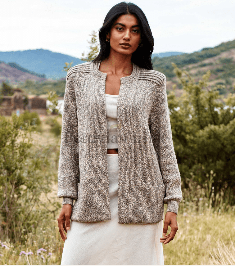Woman wearing a textured cardigan over a white outfit in a natural setting with mountains and greenery.